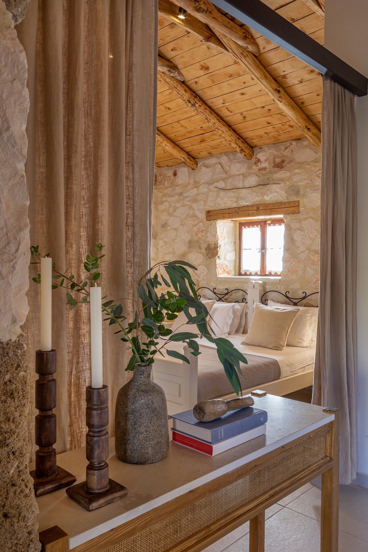 Studio bedroom seen past a linen curtain, with stone walls and a wooden roof